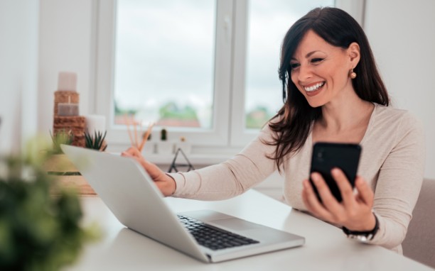 Uma mulher sorri enquanto olha para a tela de um computador enquanto segura um celular com a mão esquerda.