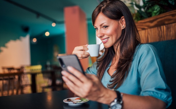 Mulher sorrindo e olhando para a tela de um celular segurado por ela com a mão esquerda, enquanto segura um xícara branca com a mão direita.