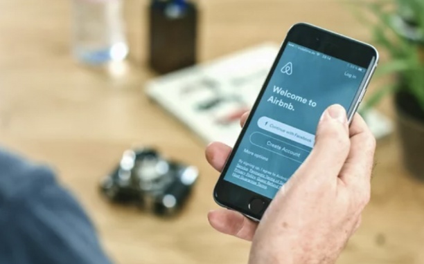 Uma mão segura um celular preto em cuja tela está aberto o aplicativo do Airbnb