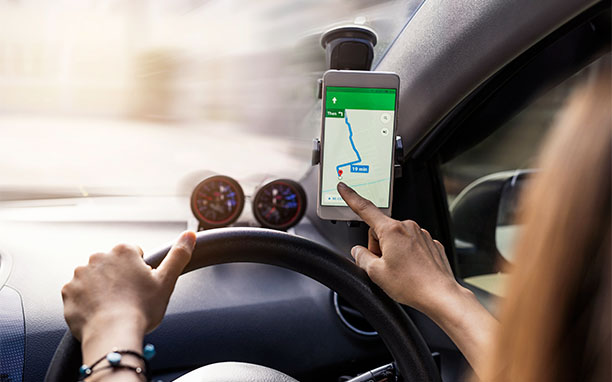 Mulher dirigindo e olhando o mapa de um app com geolocalização, em um celular que está fixado em um suporte no vidro do veículo.