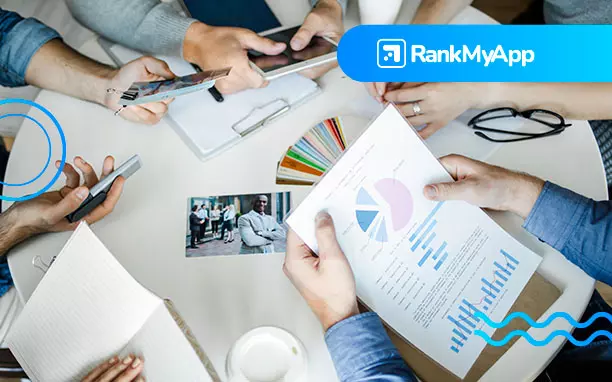 Imagem mostra braços de pessoas em volta de uma mesa, indicando que estçao em reunião. Elas seguram papéis com gráficos coloridos, celulares e em cima da mesa há mais documentos de trabalho.